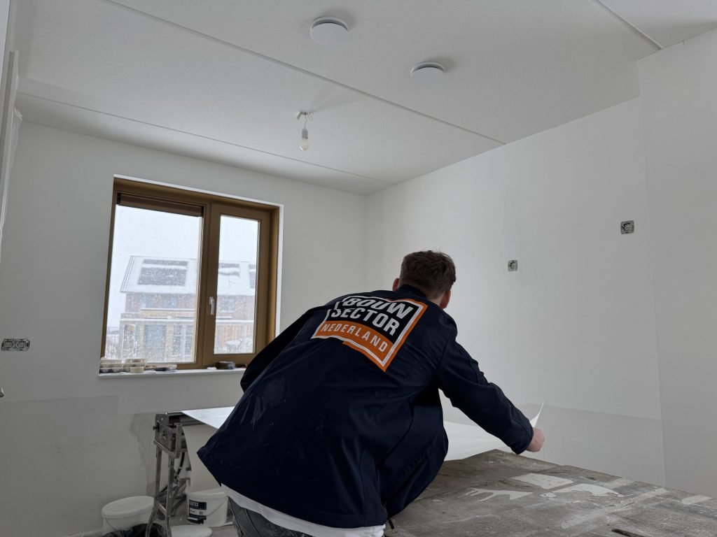 Vakman van Bouwsector Nederland in werkjas legt een baan behang op de plaktafel bij het raam