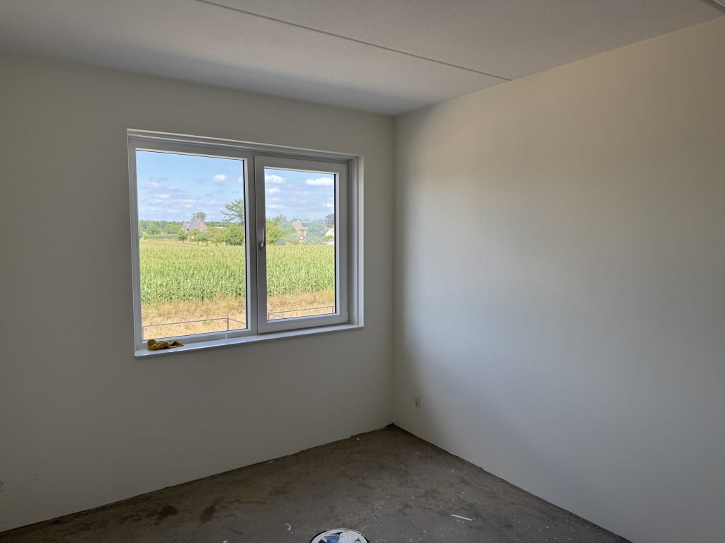 Lege nieuwbouwkamer met gladde, lichte wanden en een raam met uitzicht op een maisveld